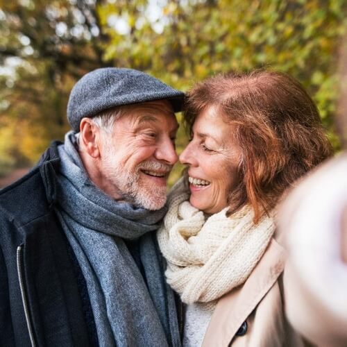 A senior couple enjoying a tender moment together outdoors, wearing cozy scarves and hats, with a backdrop of autumn leaves, symbolizing companionship and the supportive services offered by a home care and senior assistance of Qualicare